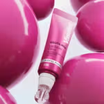 A pink tube of Vaseline Gluta‑Hya Lip Serum with a glossy applicator, surrounded by large, shiny pink spherical shapes on a soft light background.