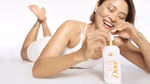 Person lying on their stomach in a white outfit, smiling while holding a bottle of Dove Glow Recharge body lotion with a pump dispenser.