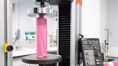 A close-up view of some lab testing equipment, showing a pink plastic bottle being compressed. The machine's control panel with various buttons is visible on the right.