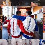 Two people walk side by side inside an Arsenal stadium concourse with their arms around each other’s shoulders, wearing red‑and‑white and blue‑and‑white jackets with ‘VARZENAL’ printed vertically on the back, with the Arsenal crest visible ahead and fans in the background