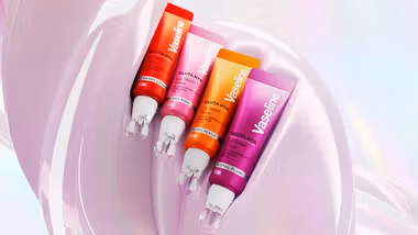 Four Vaseline Gluta Hya lip serum tubes in cherry crush, rosy pink, juicy peach and royal plum displayed against a glossy pink abstract background.