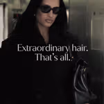 A person wearing a dark coat and sunglasses stands indoors holding a black handbag, with long dark hair visible. Overlaid text reads: “Extraordinary hair. That’s all.”
