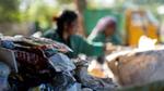 A close-up of a pile of discarded plastic packaging, with two workers sorting waste in the blurred background.