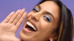 A woman with long dark hair and blue eye makeup smiles brightly against a purple background. She holds one hand up beside her face, showcasing her white teeth and happy expression.