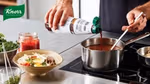 A person is cooking using an induction hob. They are stirring a pan with one hand while pouring a bottle of Knorr-branded product into the pot. Additional bowls of ingredients are arranged on the worktop.