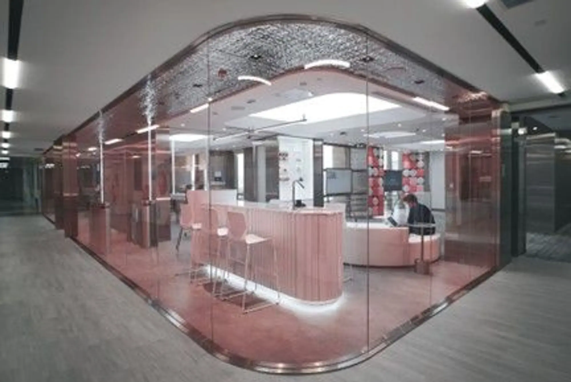 The interior of an office building, viewed from a corridor looking into a meeting room with various seating options and large TV screens. A person is sitting and working on a laptop