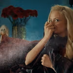 A person seated on a sofa holds a blanket close while leaning forward to smell a cloud of fine mist or spray that is dispersing in the air. A vase filled with red flowers is visible in the background against a blue wall.