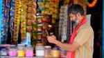 A shopkeeper wearing a striped shirt and a red scarf around his neck is using a smartphone while standing in front of a colourful shop display. The shop is filled with a variety of products, including packets hanging on the wall and jars of goods on the counter.