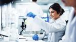 A scientist in a lab coat smiles while examining a flask of light green liquid at her workstation