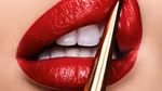 Extreme close-up of glossy red lips with white teeth, applying lipstick using a gold applicator.