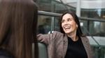 Two individuals are seated and conversing in a modern office space with glass railings and large windows. One person wears a plaid blazer over a black top and is smiling, while the other has long brown hair and is side on.