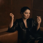 Woman sat on a brown couch dressed in black with a beauty product in her hand.