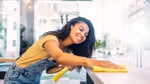 A young woman in overalls smiles while cleaning a surface with a yellow cloth inside a brightly lit room with large windows.