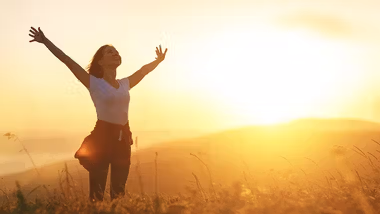 A person standing in a field with her arms raised. The sun is setting behind them casting a bright orange glow across the countryside.