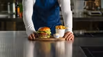 A person wearing a blue apron and leaning on a kitchen worktop. In front of them is a burger, a bowl of chips and a small serving of Hellmann’s mayonnaise, all arranged on a wooden chopping board.