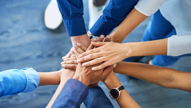 A group of people placing their hands in the centre of a circle in a pile. All are wearing clothes in shades of blue.