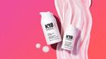 Two products from the K18 range – both white plastic bottles – lying on a bright pink background with a swirl of he product smeared on it.