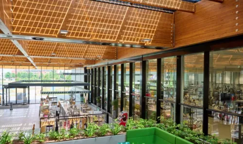 The interior of a bright and airy office building, with a foyer to the left featuring a canteen and various plants. To the right, behind glass panels, is a large office space.