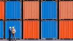 Two individuals in a shipping yard standing in front of blue and orange cargo containers. One person is wearing a white hard hat and a formal suit, and the other person is wearing a yellow hard hat, a safety vest and more casual clothing.