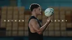 In the foreground, Nathan Cleary is spinning a rugby ball on his finger, with His name written on either side of him. In the background are rows of yellow stadium chairs.