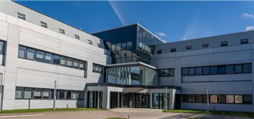 A low-level office building with a central main entrance, built predominantly from glass, with two grey wings extending to the left and right.