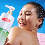 A person stands under splashing water against a bright blue background while holding a large bottle of Lifebuoy Anti Acne body wash in the foreground. The bottle has white and green packaging and features aloe vera imagery.
