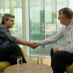 Two individuals sit facing each other in modern yellow high‑back chairs inside a bright, glass‑walled office space.