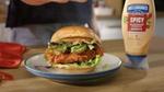 Close-up of a crispy fried chicken sandwich on a plate, layered with lettuce, pickles, and avocado slices inside a soft bun. A bottle of Hellmann’s Spicy Mayonnaise Dressing is placed to the right, and a red napkin is partially visible on the left. The setting appears to be a kitchen countertop with warm lighting.