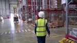 Female warehouse worker in high-visibility vest and hard hat walking towards some boxes on shelves