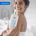 Person holding Dove Exfoliate Away body wash bottle near shoulder in a bright bathroom setting. The image is tagged ‘Geography’.