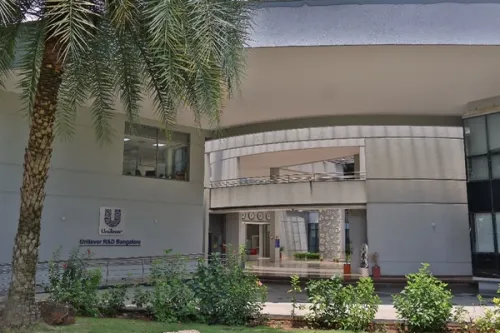 The entrance to an office building leading to a foyer. In the foreground is a garden with flowering plants and a palm tree. A sign on an exterior wall displays the Unilever logo together with the text ‘Unilever R&D Bangalore’